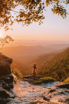 a woman standing on top of a cliff next to a river in the mountains at sunset