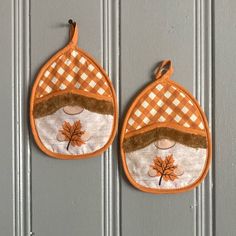 two orange and white pot holders hanging on a gray wall with an oak leaf design