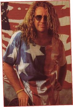 a man with long curly hair and sunglasses standing in front of an american flag background