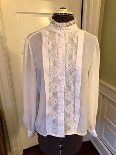 "This is an easy care yet professional blouse. This blouse is made of 100% polyester crepe and lace. There are no shoulder pads. It features a band collar with lace, front pearl button closure with vertical lace and a one inch tuck on either side. There are long sleeves with button cuffs, and a front pearl button closure. This garment is clean with no stains, snags, or odors. Measurements to guide you: Bust: 38\" Waist: 40\" Sleeve: 24\" Length from top of shoulder to hem: 26\" Decade: 70s Fabri Elegant Formal Tops With Lace Sleeves, Elegant Lace Blouse For Work, Lace Blouse With Lace Sleeves For Workwear, Elegant Lace Top For Workwear, Elegant Lace Blouse For Daywear, Elegant Lace Top For Day, Elegant Lace Work Blouse For Daywear, Feminine Blouse With Ruffled Collar For Formal Occasions, Classic Lace Tops With Lace Sleeves