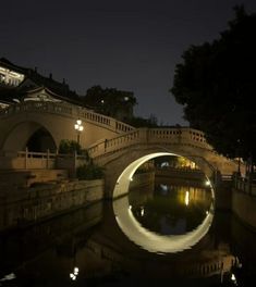 an arched bridge over a river at night with lights reflecting on the water and buildings in the background