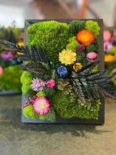 an arrangement of flowers and greenery is displayed in a square frame on a table