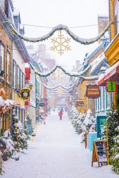 a snowy street with shops and people walking down it in the distance is decorated with christmas lights