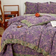 a bed covered in purple and green bedspreads with an orange rose on it