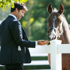 a man standing next to a brown horse near a white fence and holding the bridle