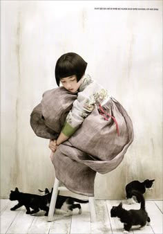 a young child is sitting on a chair with several cats around her and holding onto the pillow