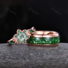 two wedding rings with green stones on them sitting on top of a gray stone surface