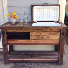 an old wooden table with ice chest and flowers on the outside patio area in front of a house