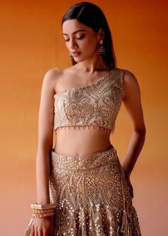 The lehenga is adorned with intricate mirrorwork and golden embellishments paired with an one-shoulder blouse in asymmetrical design, completed with mirrorwork detailing. Glamorous Saree-style Lehenga With Mirror Work, Glamorous Gold Choli With Mirror Work, Glamorous Festive Choli With Mirror Work, Party Wear Pre-draped Saree With Gota Work For Reception, Bollywood Style Lehenga With Mirror Work For Evening, Glamorous Choli With Mirror Work For Diwali, Glamorous Navratri Sharara With Mirror Work, Glamorous Designer Pre-draped Saree With Mirror Work, Glamorous Mirror Work Sharara For Navratri