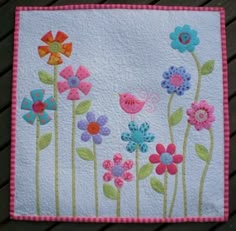 a quilted wall hanging with flowers and birds in the middle, on a wooden table