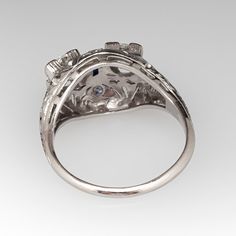 This Toi et Moi, circa 1920s, diamond ring features a pierced design with engraved details and is accented with two (2) old European cut diamonds set into four-prong settings, four (4), bead set, round single cut diamonds and one (1), bezel set, rectangular French cut synthetic blue sapphire. The ring measures 12.5mm at the top, rises 7.5mm above the finger, tapering to 1.7mm wide and 1.2mm thick at the base of the shank. It is currently a size 8. Antique Platinum Three Stone Diamond Ring, Antique Three Stone Round Diamond Ring, Antique Platinum Three Stone Rings, Antique Three Stone Platinum Rings, Heirloom Three Stone Platinum Ring, Heirloom Platinum Three Stone Ring, Heirloom Platinum Three-stone Ring, Heirloom Three-stone Platinum Ring, Art Deco Brilliant Cut Diamond Ring Collectible