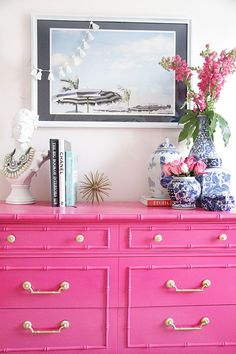 a pink dresser with flowers and vases on top, in front of a painting