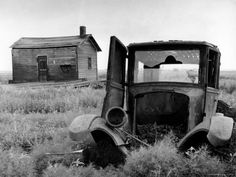 an old truck sitting in the middle of a field next to two wooden buildings and one is broken down