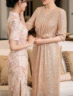 two women standing next to each other in front of a white couch wearing gold dresses