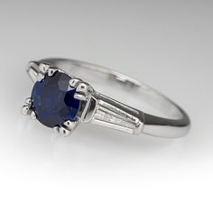 The ring is centered with one (1) round mixed cut natural sapphire set into a four-split prong setting. The shoulders of the ring are each accented with one (1), channel set, tapered baguette cut diamond. The ring measures 6.7mm at the top, rises 7.3mm above the finger, tapering to 1.8mm wide and 1.2mm thick at the base of the shank. The ring is currently a size 5.25. Timeless Sapphire Baguette Cut Ring With Prong Setting, Blue Sapphire Ring With Channel Set Baguette Cut, Baguette Cut Sapphire Platinum Ring, Platinum Sapphire Ring With Baguette Cut And Prong Setting, Silver Sapphire Ring With Prong Setting, Baguette Cut, Round Sapphire Ring, Blue Sapphire Engagement Ring, Round Sapphire, Sapphire Engagement Ring Blue