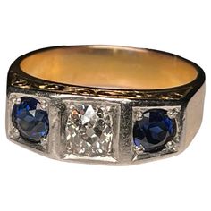 This is an Art Deco 14K Yellow Gold, Diamond and Sapphire Garter Ring. It depicts a gold ring with a top divided by two small squares at the sides and one larger one in the center. The center one is embellished by an old European brilliant diamond ( Weight- .75 carats; Measurements- 5.10 x 5.0 x 2.94 mm; Color- G; Clarity- SI-2; Type- Kimberly) and the side ones by two sapphires each one is weighing 0.5 carats. The ring is also decorated with an engraved branch of scrolls at the front and back o Formal Rectangular Sapphire Ring Fine Jewelry, 14k Yellow Gold Square Cut Diamond Ring, Formal Rectangular Sapphire Ring, 14k Gold Jewelry With Diamond Accents And Square Cut, White Gold Sapphire Ring Stamped 14k, Rectangular Brilliant Cut Sapphire Ring For Formal Occasions, Rectangular Platinum Ring For Formal Occasions, Heirloom Yellow Gold Three Stone Ring, Antique Three Stone Gold Jewelry