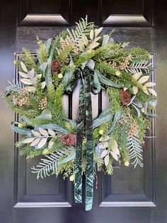 a green wreath with pine cones and evergreens hanging on a black front door frame