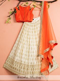 A three-piece ivory and orange mirror lehenga set from the Anisha Shetty collection. This gorgeous ivory georgette lehenga is paired with an orange cold shoulder rawsilk blouse. The ivory lehenga is enhanced with heavy sequence work all over with scalloped edging. The orange blouse has a beautiful zardozi and mirror work embroidery. This outfit is completed with an orange dupatta in net material with zari, sequence and mirror work border. Orange Choli With Mirror Work For Navratri, Orange Mirror Work Lehenga For Festive Occasions, Orange Lehenga With Mirror Work In Traditional Drape, Orange Resham Embroidery Choli In Georgette, Anarkali Style Orange Lehenga With Mirror Work, Orange Georgette Choli For Reception, Orange Mirror Work Dupatta For Reception, Off White Georgette Lehenga For Festivals, Festive Orange Georgette Choli