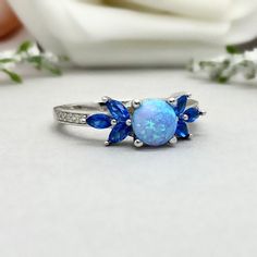 This beautiful ring is made from genuine 925 sterling silver with rhodium plating. Ring details- -The Main stone is a round cut 6mm Lab Created Light Blue Opal Stone -Side stones are 4mm by 2mm Lab Blue Sapphire Marquise and 1mm Round simulated diamonds -Ring is casted in solid 925 sterling silver with rhodium plating (yellow gold and rose gold plated also available, please check the drop down menu for more options) -The Total face height of the ring measures 7mms and the band width measures 1.5 Blue Cubic Zirconia Round Band Jewelry, Blue Cubic Zirconia Promise Jewelry, Blue Round Band Jewelry For Promise, Blue Round Band Promise Jewelry, Blue Cluster Ring With Prong Setting For Promise, Blue Halo Setting Birthstone Ring For Anniversary, Blue Round Cut Promise Ring, Blue Cubic Zirconia Crystal Ring, Blue Cubic Zirconia Rings With Round Cut