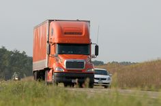 an orange semi truck driving down a road next to a white car in the grass