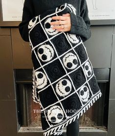 a woman wearing a black and white crocheted blanket with skulls on it, standing in front of a fireplace