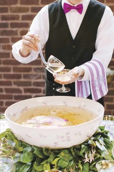 a man in a vest pouring wine into a bowl on top of a table with greenery