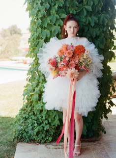 a woman holding a bouquet of flowers in front of a green bush with pink ribbon