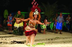 a woman in a hula skirt dancing on the beach with other people behind her