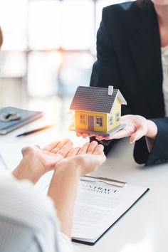 two people sitting at a table with a small model house on their hands and papers in front of them