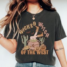 Stir up some Halloween fun with our "Wicked Witch of the West" T-shirt! This enchanting design features a bubbling cauldron with legs in cowgirl boots sticking out, surrounded by whimsical decorations like cacti, a broomstick, and jack-o-lanterns. Perfect for adding a touch of spooky Western charm to your fall wardrobe, this shirt is ideal for Halloween parties, casual outings, or cozying up with a spellbook. Made from soft, high-quality fabric, it ensures comfort and style as you embrace your w Witchy Short Sleeve T-shirt With Graphic Print, Witchy Halloween T-shirt With Letter Print, Halloween Cartoon Print Relaxed Fit Tops, Witchy Cotton Tops With Letter Print, Witchy Cotton Crew Neck Tops, Witchy Graphic Print Crew Neck Tops, Witchy Letter Print T-shirt For Fall, Witchy Halloween T-shirt With Graphic Print, Fall Witchy Crew Neck T-shirt