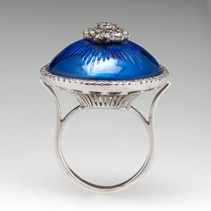 This regal white gold cocktail ring features an engraved yellow gold dome design, topped with blue enamel. The center of the dome is prong set with a cluster of nine (9) round brilliant cut diamonds. The dome is surrounded by a halo, scallop set with sixty (60) round brilliant cut diamonds. The ring measures 24.3mm at the top, rises 15.8mm above the finger, tapering to 2.6mm wide and 1.3mm thick at the base of the shank. It currently fits a size 6. Elegant Blue Cabochon Dome Ring, Art Deco Enamel Rings For Formal Occasions, Luxury White Gold Enamel Oval Ring, Blue Enamel Ring With Polished Finish, Blue Enamel Polished Round Ring, Blue Polished Enamel Ring, Elegant Formal Enamel Ring, Classic Blue Domed Ring, White Gold Luxury Enamel Ring For Formal Occasions