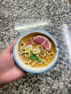 a hand holding a bowl of ramen with meat and noodles in it on a marble countertop