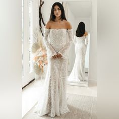 a woman standing in front of a mirror wearing a white wedding dress with off the shoulder sleeves