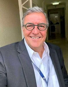 an older man wearing glasses and a white shirt smiles at the camera while standing in front of a hallway