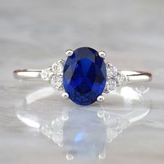 ♥ Beautifully handcrafted Blue Sapphire ring that features 1.5-carat classic oval cut Sapphire gemstone with brilliant-cut Diamond accents (5A grade CZ) in solid Sterling Silver. ♥ This ring makes the perfect engagement or anniversary gift for her that will be treasured forever! ♥ Blue Sapphire is traditionally associated with love and purity. It's said to bring prosperity and attract gifts of all kinds. Metal: Solid 925 Sterling Silver Gemstone: Blue Sapphire Side Stones: Diamond simulants (5A Oval Blue Birthstone Ring With Center Stone, Blue Oval Birthstone Ring In Fine Jewelry Style, Blue Oval Diamond Ring, Oval Rings With Side Stones For Gift, Oval Diamond Ring With Side Stones For Promise, Blue Oval Birthstone Ring For Anniversary, Blue Rings With Side Stones And Round Cut, Oval Diamond Promise Ring With Side Stones, Oval Blue Birthstone Ring For Anniversary