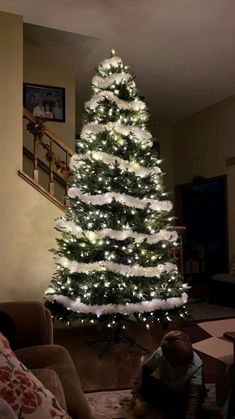 a white christmas tree is lit up in the living room with a toddler playing under it