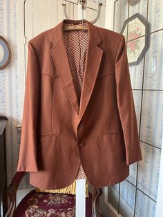 This jacket from Mister Jack's Men's Wear Ltd. is made from a wool blend in rust brown. It has the original buttons in the front and on the cuffs, two front pockets and two inside pockets, and it's fully lined with brown, beige and orange print lining. It has western styling on the front and back yokes and pockets. The measurements, taken with the coat lying flat, are: shoulder to shoulder, 18 inches; armpit to armpit, 23 inches; sleeves, 27 inches; length, 36  inches; bottom edge, 34 inches. There is a small light spot on the right lapel (see photo), but otherwise the coat is in very good condition. Brown Flat Front Suit For Fall, Brown Flat Front Suits For Fall, Brown Tweed Jacket With Single Button, Brown Fitted Outerwear With Single Button, Fitted Brown Outerwear With Single Button, Brown Single Breasted Suits For Winter, Brown Single Button Long Sleeve Tweed Jacket, Brown Long Sleeve Suit For Fall, Brown Single-breasted Suit For Winter