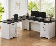 a desk with two computers on it in front of a window and wood flooring