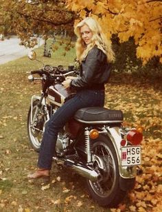 a woman sitting on top of a motorcycle next to trees with leaves all over the ground