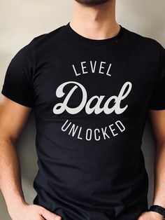 "The \"Dad Level Unlocked\" t-shirt is a fun and playful way for dads and soon-to-be dads to show off their fatherhood achievements. The text is a nod to the popular gaming culture phrase \"level unlocked,\" and highlights the new level of responsibility and joy that comes with becoming a dad. This t-shirt makes a great gift for Father's Day, birthday, or for a baby shower/baby announcement!  Bella + Canvas Brand Shirt ▶Unisex Adult Sizing ▶See Our Size Chart For Proper Sizing ▶For an oversized look, go 1-2 sizes up! ▶Rolled sleeves are for styling purposes only ▶Props used in photos are not included with the purchase ▶Please note that the colors of the design on the T-shirt may      appear slightly different due to variations in monitor settings. ✈ PROCESSING & SHIPPING ✈ Processing Time: Black T-shirt For Father's Day Parenting, Dad Tshirt Ideas, Dad Pregnancy Announcement, Expectant Father, First Time Dad, Father Shirts, Diy Shirts, Pregnancy Announcement Shirt