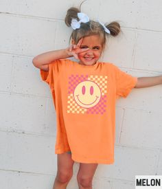 Our retro smiley face graphic tee features a happy face and checkered background design. This tshirt pairs perfectly with the adult, youth, and toddler girls versions (linked below). ✩ ✩ 𝗧𝗼𝗱𝗱𝗹𝗲𝗿 𝗕𝗲𝗹𝗹𝗮 𝗖𝗮𝗻𝘃𝗮𝘀 𝗧𝗲𝗲: https://starrieapparel.etsy.com/listing/1703012396 ✩ 𝗞𝗶𝗱𝘀 𝗕𝗲𝗹𝗹𝗮 𝗖𝗮𝗻𝘃𝗮𝘀 𝗧𝗲𝗲: https://starrieapparel.etsy.com/listing/1702998672 ✩ 𝗞𝗶𝗱𝘀 𝗖𝗼𝗺𝗳𝗼𝗿𝘁 𝗖𝗼𝗹𝗼𝗿𝘀 𝗧𝗲𝗲: https://starrieapparel.etsy.com/listing/1728079245 ✩ 𝗔𝗱𝘂𝗹𝘁 𝗕𝗲𝗹𝗹𝗮 Cheap Funny T-shirt With Smiley Face, Affordable Short Sleeve T-shirt With Smiley Face, Cheap Smiley Face Short Sleeve T-shirt, Fun Smiley Face Short Sleeve T-shirt, Cute Orange T-shirt With Graphic Print, Pink Short Sleeve T-shirt With Smiley Face, Cute Smiley Face Short Sleeve T-shirt, Cute Short Sleeve Top With Smiley Face, Trendy Short Sleeve T-shirt With Smiley Face