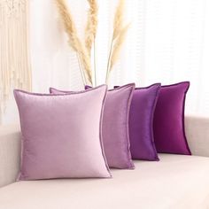 four purple pillows sitting on top of a white couch next to a vase with dry grass in it