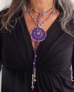 a woman with grey hair wearing a purple beaded necklace and black top, standing in front of a white wall