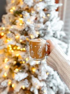 a woman holding a cup of coffee in front of a christmas tree with lights on it