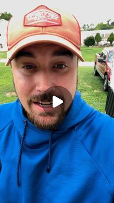 a man with a beard wearing a blue hoodie and a baseball cap is standing in front of a house