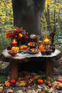 a table with candles and fruit on it in front of a large tree surrounded by leaves