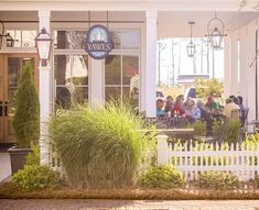 a group of people sitting outside of a restaurant next to a white picket fence and trees