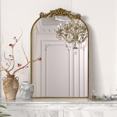 a white mantle topped with a mirror and vase filled with flowers next to a gold framed mirror