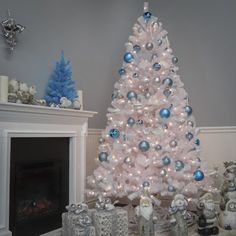 a white christmas tree decorated with blue and silver ornaments in front of a fire place