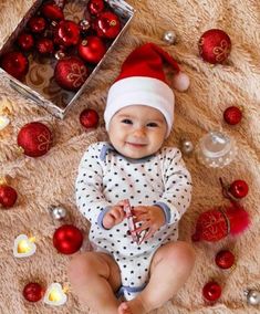 a baby wearing a santa hat and sitting on a rug surrounded by christmas ornaments, candles and balls